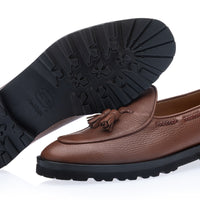 A pair of brown pebbled leather tassel loafers shown on a white background, with one shoe standing upright to display the side and sole, and the other lying flat to show the treaded black outsole. | Image 2