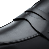 Close-up of a black leather penny loafer showing the smooth textured leather, decorative strap, and fine stitching against a white background. | Image 5