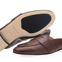 A pair of brown leather slip-on mule loafers shown on a white background, with one shoe turned to display the beige insole and black outsole. | Image 2